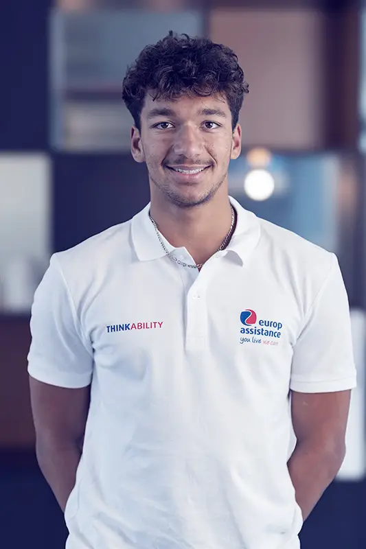 Person wearing a white Europ Assistance polo shirt with the slogan 'You live we care' and the word 'THINKABILITY,' standing indoors.
