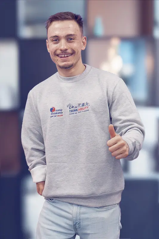 A smiling young man with short hair and a trimmed beard looks directly at the camera. He’s wearing a light gray sweatshirt featuring two logos—“Europ Assistance – you live we care” and “ThinkAbility – Look up, see beyond”—and he gives a thumbs-up with his right hand. The background is softly blurred to keep the focus on him.