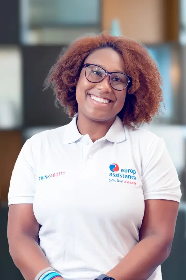 Person wearing a white Europ Assistance polo shirt with the slogan 'You live we care' and the word 'THINKABILITY,' standing indoors.