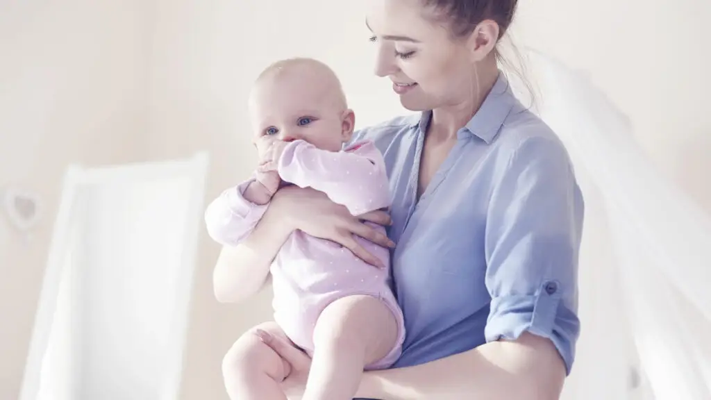 A person holding a baby dressed in a light-colored onesie while standing indoors in a softly lit room with light-colored walls and a sheer canopy in the background